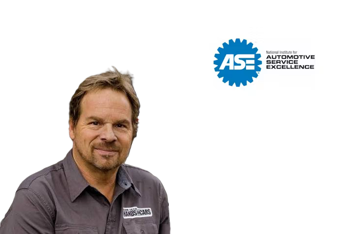 Professional male automotive technician wearing gray work shirt with embroidered logo, smiling at camera with ASE certification badge displayed
