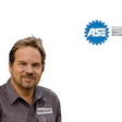 Professional male automotive technician wearing gray work shirt with embroidered logo, smiling at camera with ASE certification badge displayed