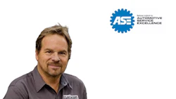 Professional male automotive technician wearing gray work shirt with embroidered logo, smiling at camera with ASE certification badge displayed