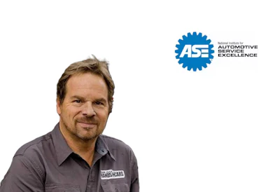 Professional male automotive technician wearing gray work shirt with embroidered logo, smiling at camera with ASE certification badge displayed