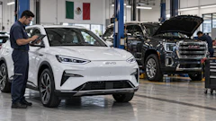 Technician inspecting white BYD electric vehicle in automotive service facility with black GMC truck and Mexican flag visible