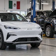 Technician inspecting white BYD electric vehicle in automotive service facility with black GMC truck and Mexican flag visible