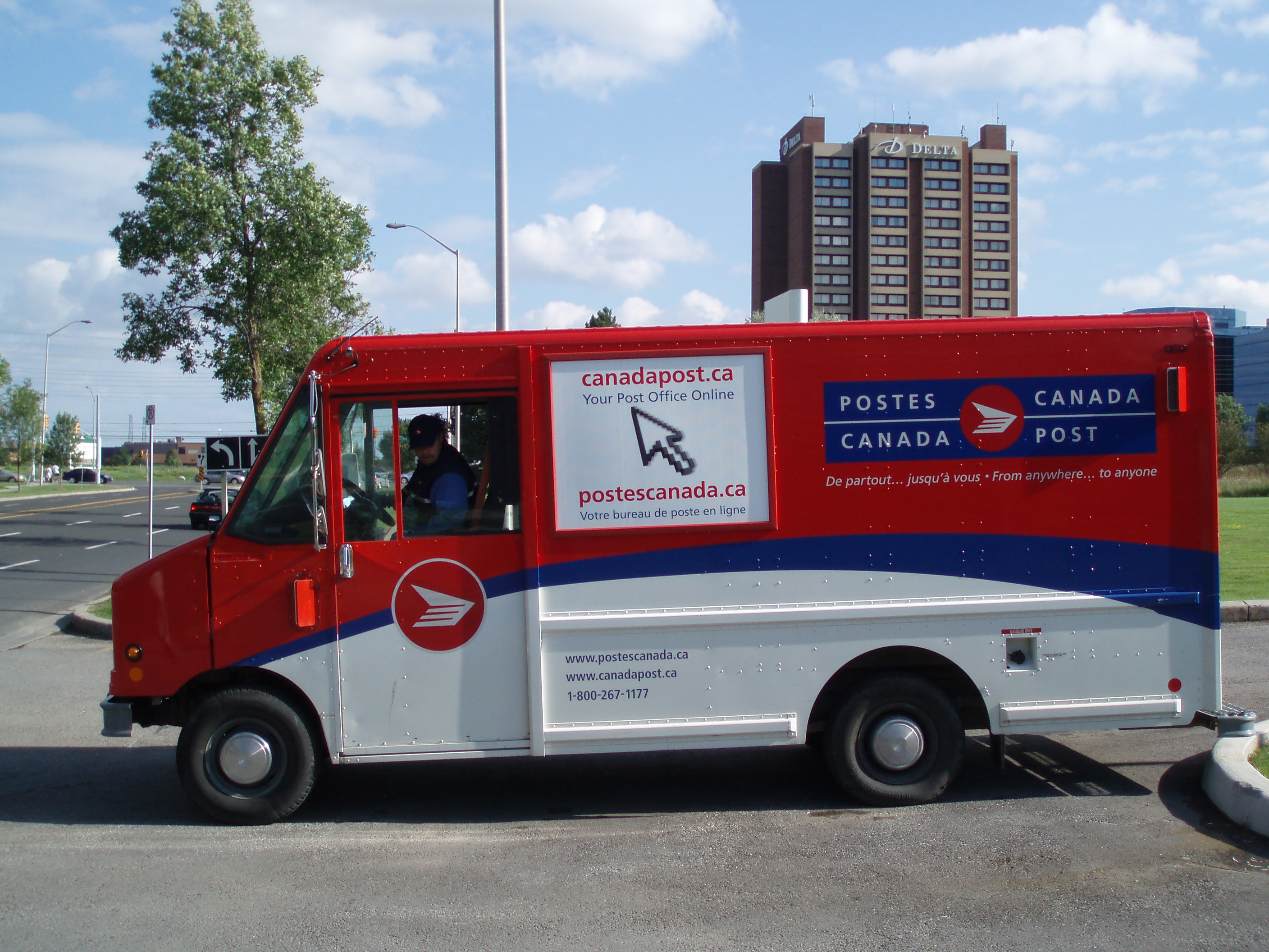 Red and white Canada Post delivery truck with postal service branding and website information on city street
