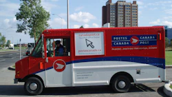 Red and white Canada Post delivery truck with postal service branding and website information on city street