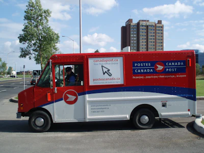 Red and white Canada Post delivery truck with postal service branding and website information on city street