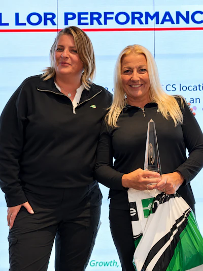 Two women at corporate event, one holding glass award, standing before branded backdrop