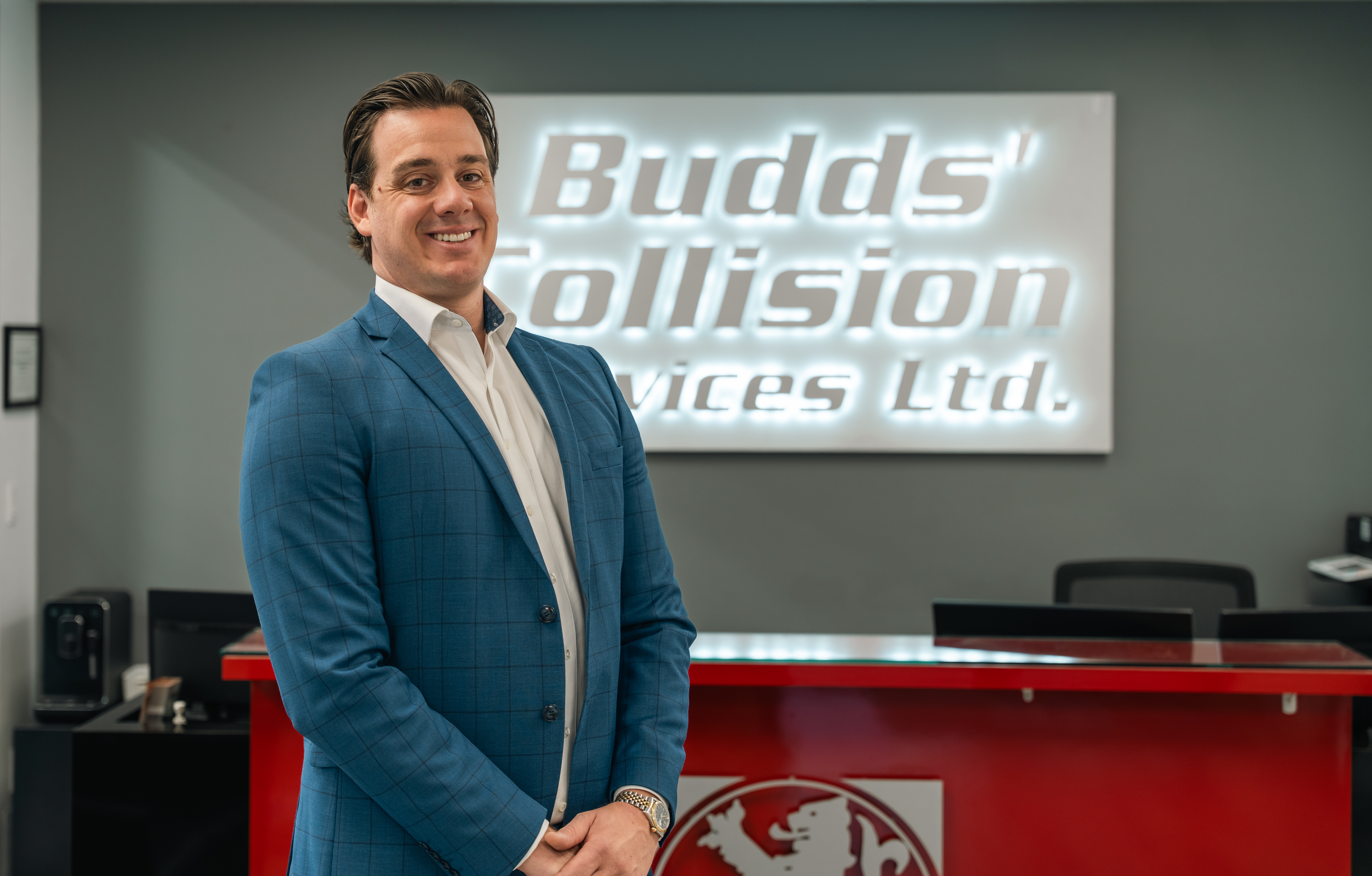 Professional man in blue blazer standing in modern office with Budds' Collision Services Ltd. signage