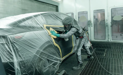 Automotive technician in protective gear spray painting a vehicle in an industrial paint booth facility