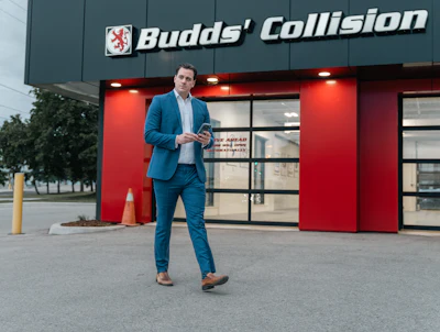 Professional man in blue suit standing outside Budds' Collision auto repair shop with red and teal storefront