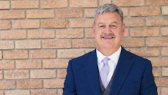 Professional headshot of a man in navy blazer and light purple tie against brick wall background