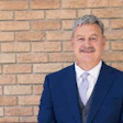 Professional headshot of a man in navy blazer and light purple tie against brick wall background