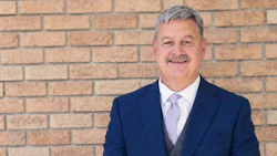 Professional headshot of a man in navy blazer and light purple tie against brick wall background