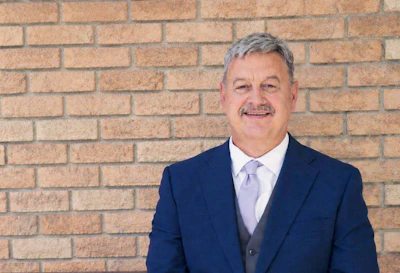 Professional headshot of a man in navy blazer and light purple tie against brick wall background