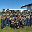 Team members in matching branded apparel pose together outdoors in front of an RV at a sporting event