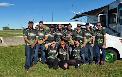 Team members in matching branded apparel pose together outdoors in front of an RV at a sporting event