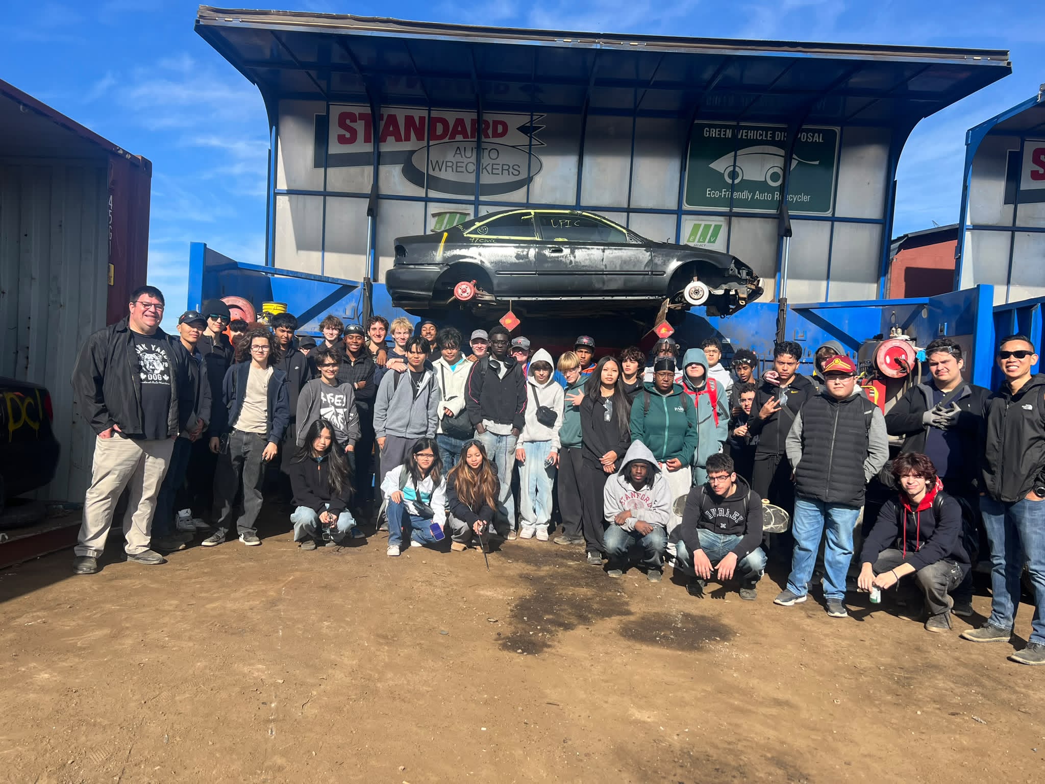 Large group of people posing together outside Standard Auto Wreckers facility with vintage car displayed above entrance