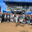 Large group of people posing together outside Standard Auto Wreckers facility with vintage car displayed above entrance