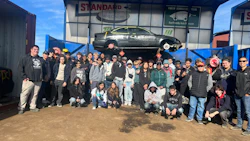 Large group of people posing together outside Standard Auto Wreckers facility with vintage car displayed above entrance