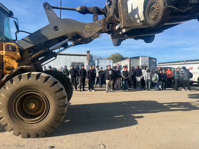 Heavy equipment operator lifts vehicle with industrial crane at scrapyard facility with crowd of spectators watching