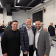 Three men smiling at a corporate event in a modern venue with attendees in the background