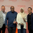 Four professionals posing together at a Simplicity branded corporate event with coral gradient backdrop