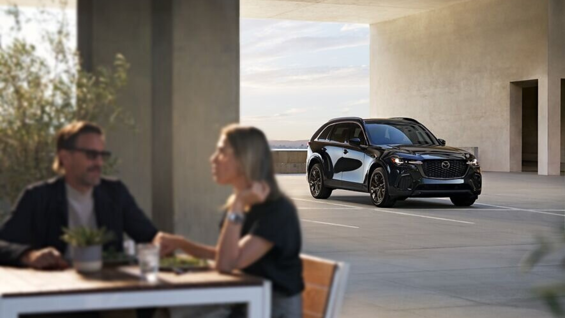 Two professionals discuss business while a sleek black Mazda SUV is displayed in a modern architectural space with floor-to-ceiling windows.