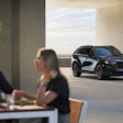 Two professionals discuss business while a sleek black Mazda SUV is displayed in a modern architectural space with floor-to-ceiling windows.