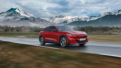 Red Ford Mustang Mach-E electric vehicle driving on wet road with snow-capped mountains in background