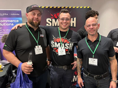 Three men wearing event badges smile together in front of a SMASH branded backdrop at a conference or networking event.