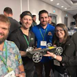 Group of five people at a tech event holding a robotic device with white wheels and wearing conference badges