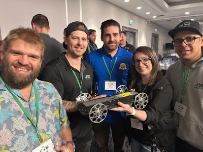 Group of five people at a tech event holding a robotic device with white wheels and wearing conference badges