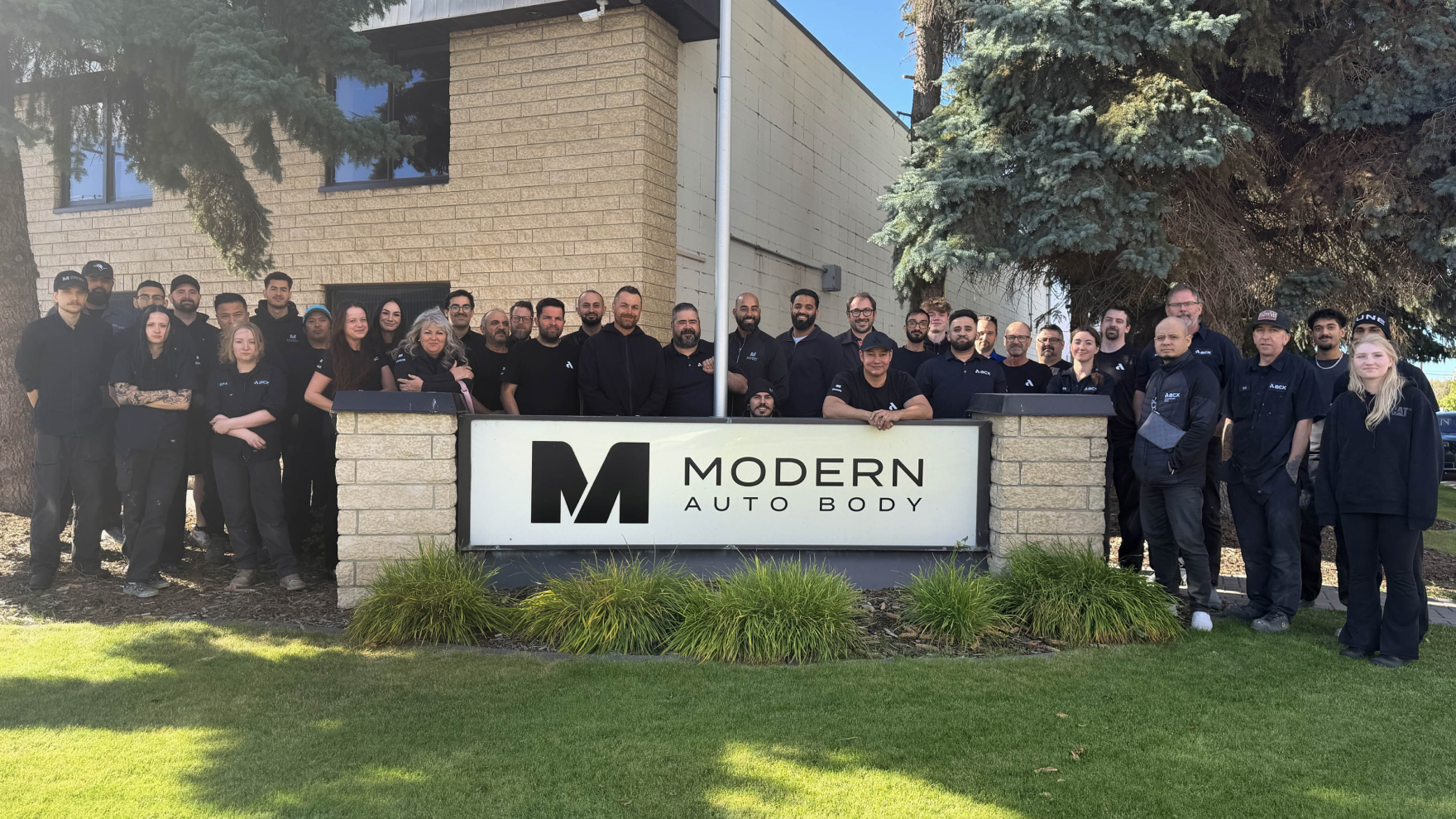 Modern Auto Body team posing together in front of company sign with brick building
