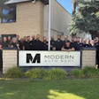 Modern Auto Body team posing together in front of company sign with brick building