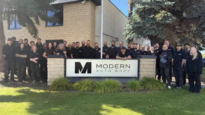 Modern Auto Body team posing together in front of company sign with brick building